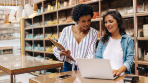 Two female colleagues standing at a table looking at a laptop.