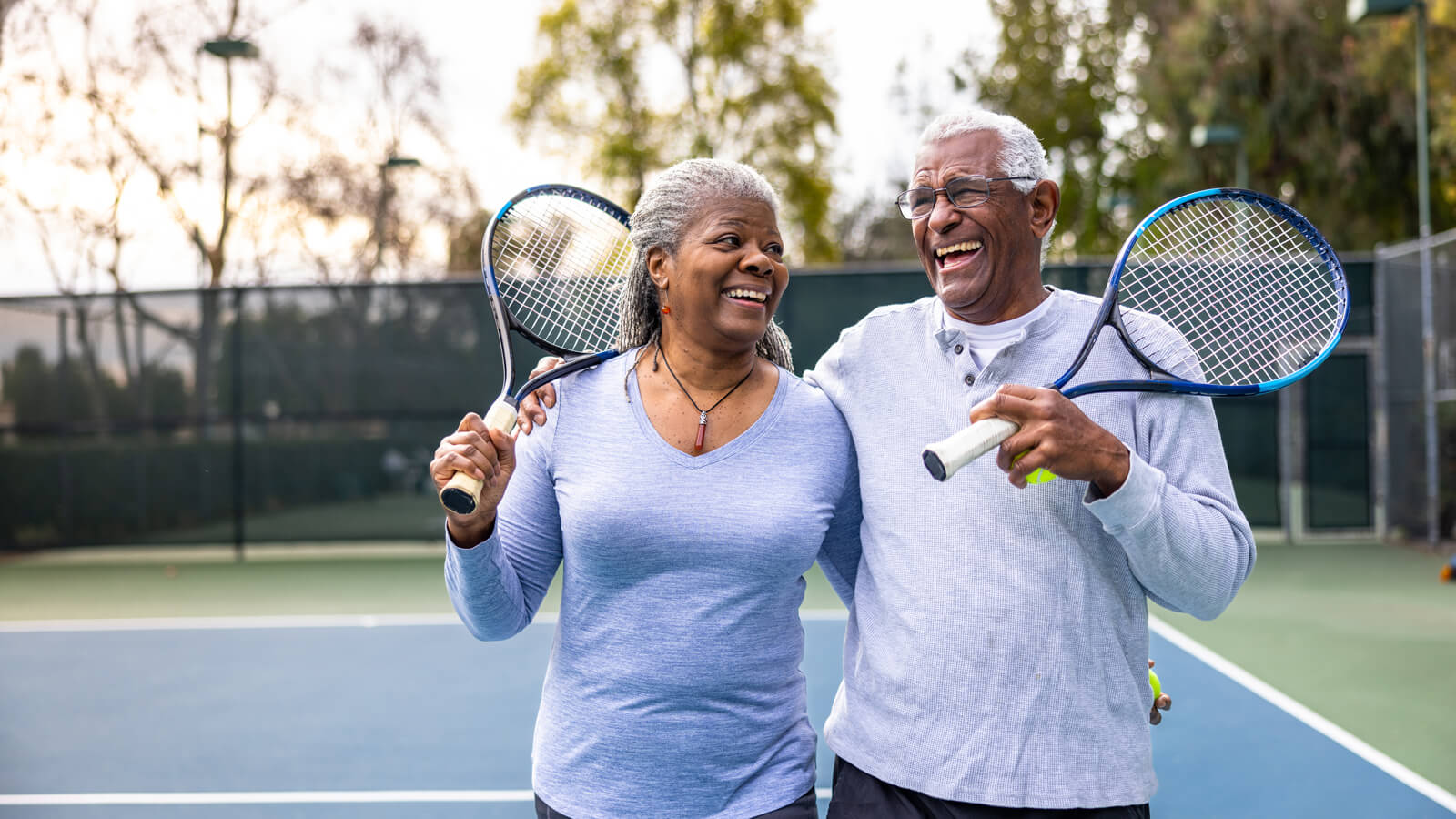 A husband and wife in their 60s walk off a paddleboard court after finishing a game with their arms around each other smiling.