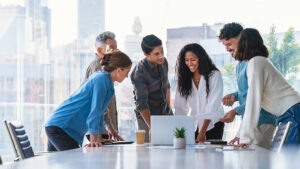 A group of colleagues in an office gathered around a table looking at a laptop.