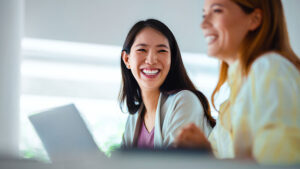 A professional woman in her 30s smiles at her coworker during a meeting in a corporate office.