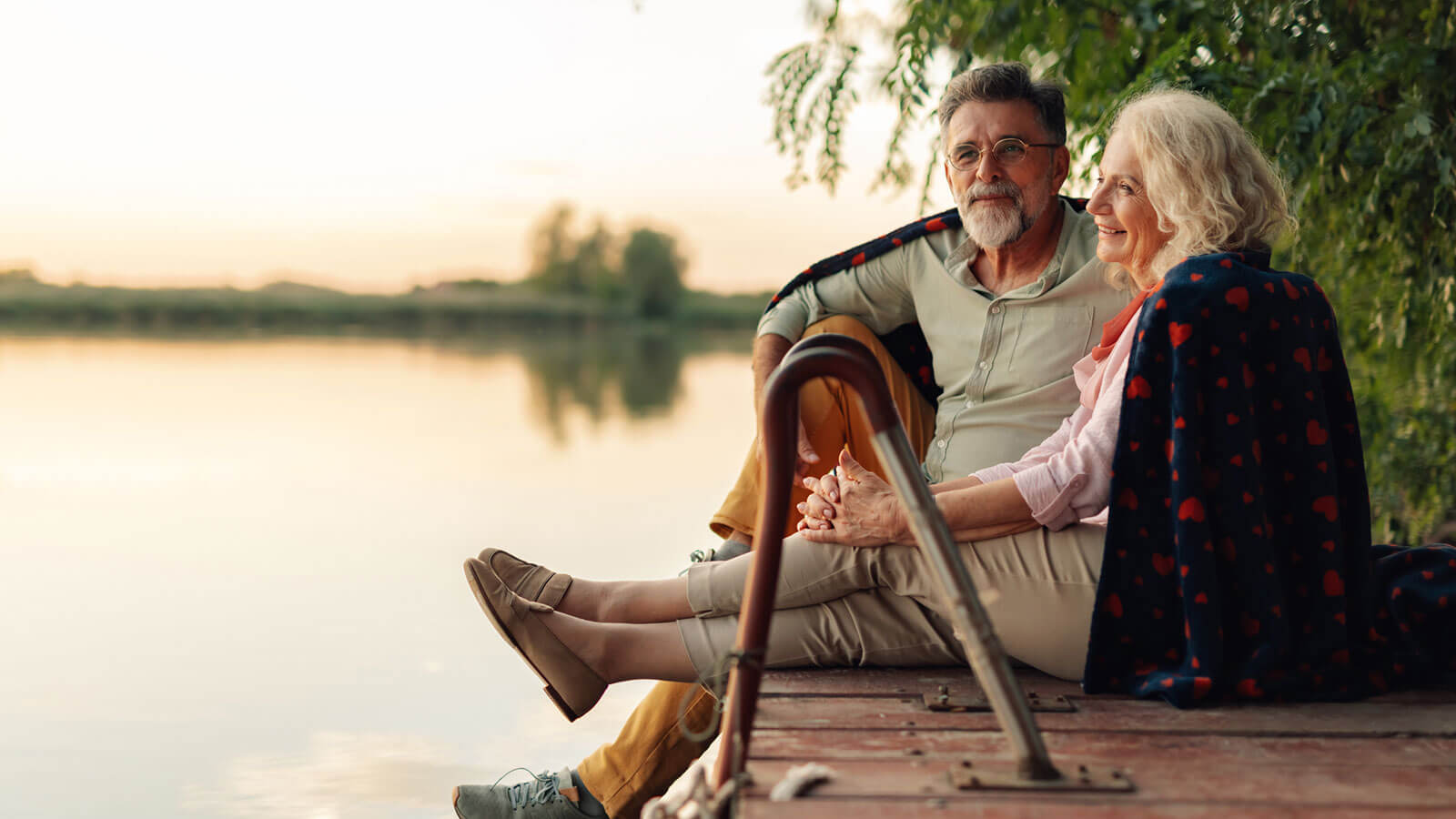A husband and wife in their late 60s are sitting happily together on a dock overlooking a lake at sunset enjoying their retirement years.
