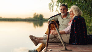 A husband and wife in their late 60s are sitting happily together on a dock overlooking a lake at sunset enjoying their retirement years.