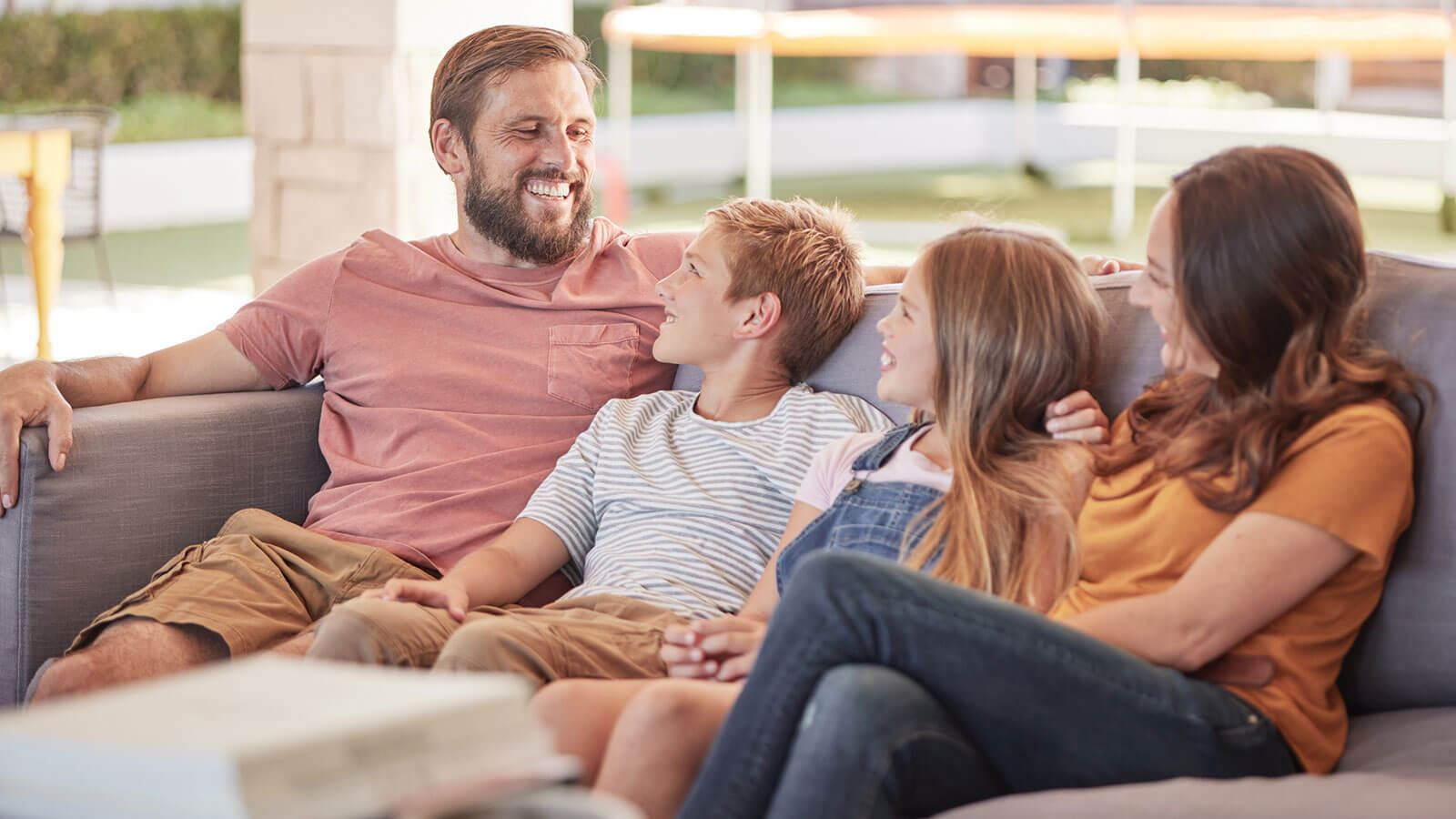 A dad and mom sit on either side of their two kids on an outdoor couch, the whole family smiling happily on a beautiful, sunny day.