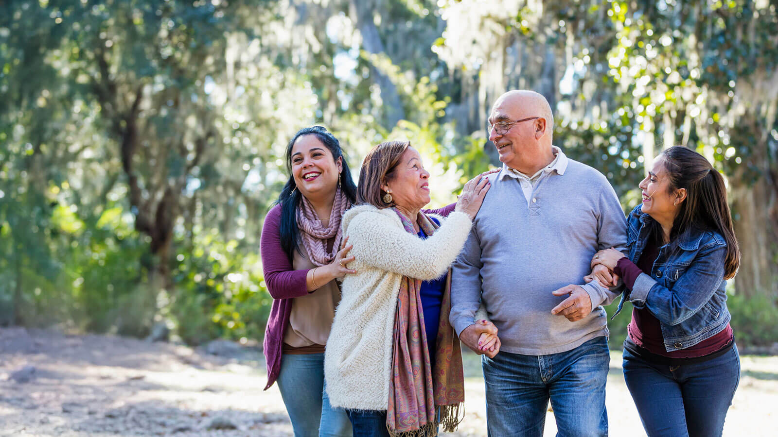 A family of four, two parents and their two adult daughters, are taking a walk on a sunny, fall day through a wooded park, laughing and having a fun afternoon together.